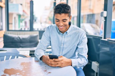 Kahve dükkanının terasında akıllı telefon kullanarak mutlu gülümseyen genç Latin adam..