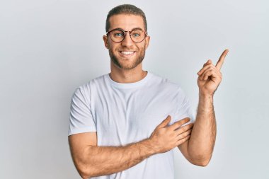Young caucasian man wearing casual clothes and glasses with a big smile on face, pointing with hand and finger to the side looking at the camera. 
