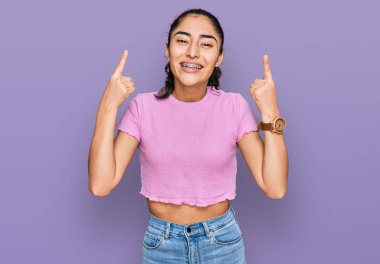 Diş teli takan, sıradan giyinen, şaşkın şaşkın gülümseyen, parmaklarıyla işaret eden ve kollarını kaldıran İspanyol genç kız.. 