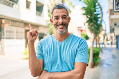 Orta yaşlı, kır saçlı, günlük kıyafetler giyen, gülümseyen, mutlu, eli ve parmağı kenarda bir adam. 