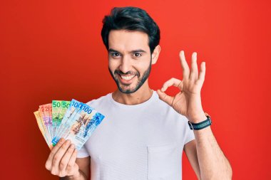 İspanyol asıllı genç adam İsviçre frangı banknotları tutuyor. Parmaklarıyla imza atıyor, gülümsüyor ve mükemmel bir sembol oluşturuyor. 