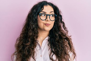 Kıvırcık saçlı, sıradan kıyafetler ve gözlük takan genç esmer kadın gülümsüyor ve başka tarafa bakıyor.. 