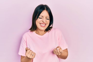 Genç İspanyol kadın, gündelik pembe tişört giyiyor. Çok mutlu ve heyecanlı. Kolları havada, gülümseyen ve başarı için bağıran kazanan bir jest yapıyor. kutlama konsepti. 