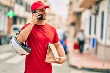 Beyaz atlı genç teslimatçı akıllı telefondan konuşuyor ve şehirde kağıt torba taşıyor..