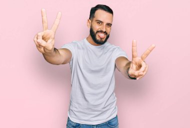 Sakallı, günlük beyaz tişörtlü, dili dışarı fırlamış bir genç zafer işareti yaparken parmaklarını gösteriyor. İki numara.. 