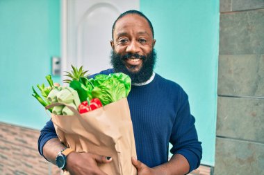 Sakallı Afrikalı Amerikalı bir adam elinde süpermarketten alınmış kağıt poşetler tutuyor.