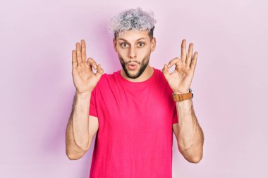 Modern boyalı saçlı, sıradan pembe tişörtlü genç bir İspanyol. Şaşırmış ve şok olmuş görünüyor. Tamam sembolünü parmaklarıyla yapıyor. çılgın ifade 