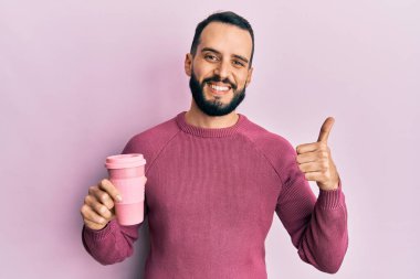 Sakallı genç adam bir fincan kahve içiyor mutlu ve pozitif gülümsüyor, başparmağıyla mükemmel ve onay işareti yapıyor. 