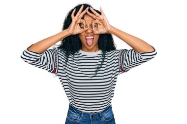 Orta yaşlı Afro-Amerikalı kadın günlük kıyafetler giyiyor dürbün gibi jest yapıyor, dili dışarı çıkarıyor, gözleri parmaklarından bakıyor. çılgın ifade. 