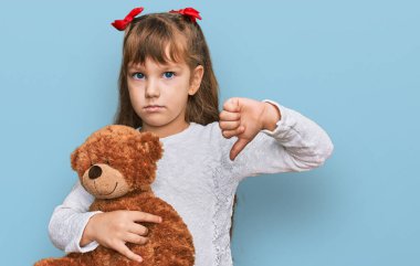 Kafkasyalı küçük kız oyuncak ayıya sarılmış kızgın yüzlü, negatif işaretli, başparmakları aşağıda, reddedilme kavramından hoşlanmadığını gösteren. 