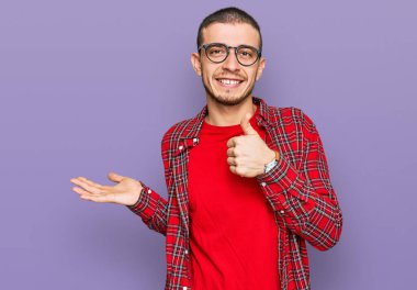 İspanyol asıllı genç adam, günlük kıyafetler giyiyor, avuç içini gösteriyor ve baş parmaklarıyla iyi bir hareket yapıyor, mutlu ve neşeli gülümsüyor. 