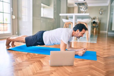 Orta yaşlı, sakallı ve esnek bir adam evde egzersiz yapıyor bilgisayardaki spor videolarına bakıyor.