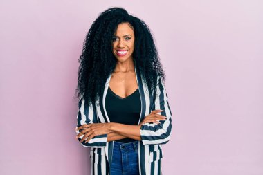 Orta yaşlı Afro-Amerikalı kadın günlük kıyafetler giyip kollarını kavuşturup kameraya bakarken gülümsüyor. pozitif kişi. 