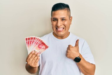 Elinde 20 İsrail şekeri tutan genç Latin adam mutlu ve olumlu gülümseyen banknotlar, mükemmel ve onay işareti yapan başparmak. 