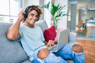 Evde dizüstü bilgisayar ve kulaklık kullanarak mutlu gülümseyen genç İspanyol adam.