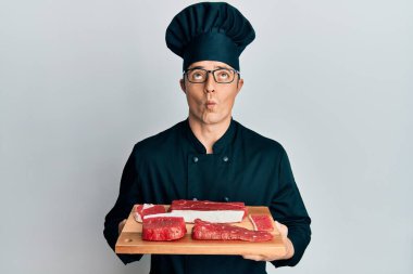Yakışıklı genç adam, şef üniforması içinde çiğ etle tahta tutuyor balık suratı yapıyor, gözleri kısık, deli ve komik.. 
