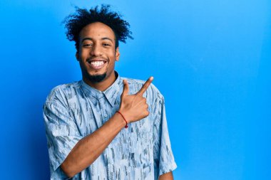 Sakallı, neşeli giyinmiş, yüzünde mutlu ve doğal bir ifadeyle, elleri ve parmaklarıyla yüzünü gösteren gülümseyen Afro-Amerikalı genç bir adam. 