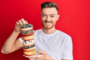 Genç kızıl saçlı adam yüzünde mutlu ve serin bir gülümsemeyle gülümseyen lezzetli renkli çörekler tutuyor. diş gösterme. 
