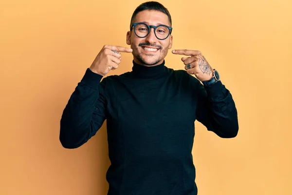 タトゥーネックのセーターと眼鏡を身に着けている入れ墨のハンサムな男は 陽気な表情と指の歯と口で指して笑っています 歯の健康の概念 — ストック写真