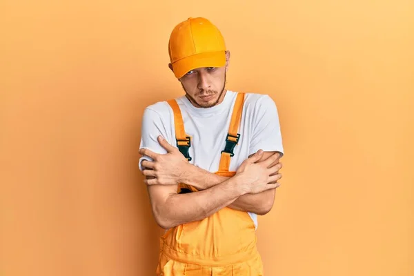 Hispanic young man wearing handyman uniform shaking and freezing for winter cold with sad and shock expression on face