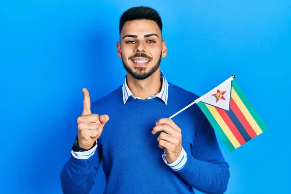 Joven Hombre Hispano Con Barba Sosteniendo Bandera Zimbabwe Sonriendo Con —  Fotos de Stock
