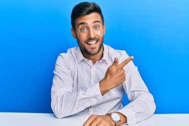 Yakışıklı İspanyol bir adam iş kıyafetleri içinde neşeli bir şekilde masada oturuyor yüzünde bir gülümseme, mutlu ve doğal bir ifadeyle elini ve parmağını yana kaldırıyor. 