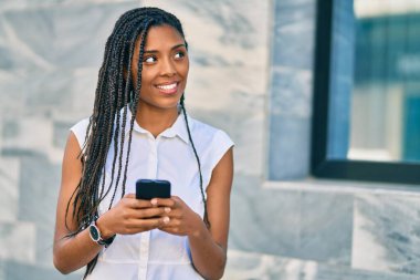 Genç Afrikalı Amerikalı kadın şehirde akıllı telefon kullanarak mutlu bir şekilde gülümsüyor..