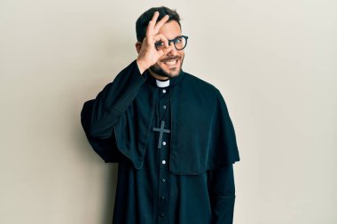 Beyaz arka plan üzerinde duran İspanyol üniformalı genç adam el hareketleriyle gülümsüyor ve mutlu yüzüyle parmaklarının arasından bakıyor.. 