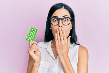 Elinde doğum kontrol hapları tutan genç ve güzel bir kadın. Ağzını eliyle kapatıyor. Şok geçiriyor ve hata yapmaktan korkuyor. şaşırmış ifade 