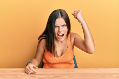 Kafkasyalı genç bir kadın, gündelik giysiler giymiş kızgın ve kızgın bir şekilde masaya oturmuş öfkeyle bağırırken yumruğunu kaldırıyor. öfke ve agresif konsept. 