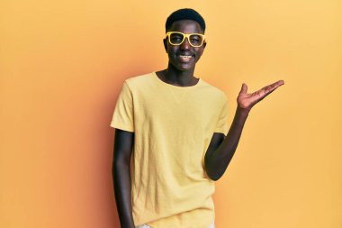 Genç Afro-Amerikan adam günlük kıyafetler ve gözlük takıyor neşeli bir şekilde gülümsüyor ve avuç içi kameraya bakıyor.. 