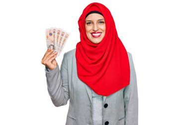 İslami tesettürlü güzel bir kadın elinde 5 kilo banknot tutarken pozitif ve mutlu görünüyor ve gülümseyerek dişlerini gösteriyor. 