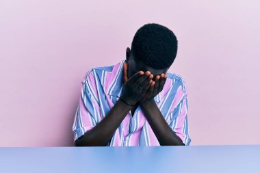 Gündelik giysiler giymiş genç Afro-Amerikan bir adam masaya oturmuş ağlarken suratını ellerle kapatıyor. depresyon kavramı. 