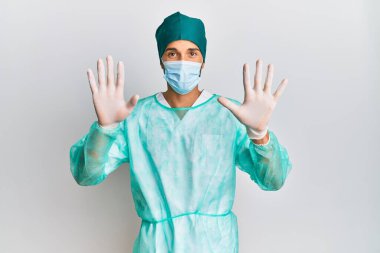 Young handsome man wearing surgeon uniform and medical mask showing and pointing up with fingers number ten while smiling confident and happy. 