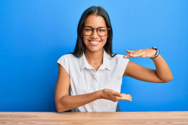 Güzel İspanyol kadın, gündelik giysiler giyip masaya oturmuş büyük ve büyük boy işaretleri göstererek el kol hareketi yapıyor. Ölçü sembolü. Kameraya bakıp gülümsüyor. ölçüm kavramı. 