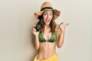 Young brunette woman holding avocado pointing thumb up to the side smiling happy with open mouth 