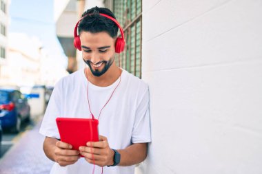 Genç Arap adam şehirde kulaklık ve dokunmatik ped kullanarak mutlu bir şekilde gülümsüyor..