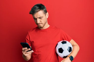 Elinde futbol topu tutan yakışıklı beyaz bir adam akıllı telefona bakıyor. Depresyonda ve endişe içinde, sinirli ve korkmuş. hüzünlü ifade. 