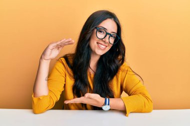 Güzel esmer, gözlüklü genç bir kadın masanın üzerinde oturmuş, büyük ve büyük beden işareti, ölçü sembolü gösteren el hareketleriyle işaret ediyor. Kameraya bakıp gülümsüyor. ölçüm kavramı. 