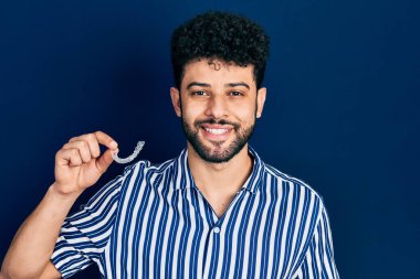 Sakallı genç Arap adam, görünmez hizalı diş telleri tutuyor. Olumlu ve mutlu görünüyor. Kendine güvenen bir gülümsemeyle dişlerini gösteriyor. 