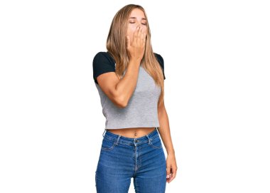 Gündelik kıyafetler giyen genç sarışın kadın ağzıyla ağzını kapatmaktan sıkılmış. Huzursuz ve uykulu.. 