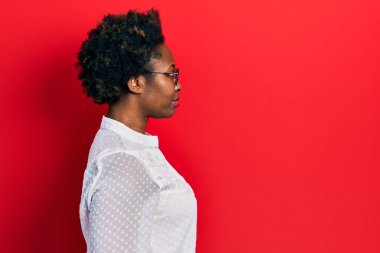 Afro-Amerikalı genç bir kadın günlük kıyafetler ve gözlük takıyor. Yan tarafa bakıyor, rahat poz veriyor, kendine güvenen bir gülümsemesi var.. 