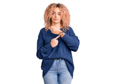 Kıvırcık saçlı, sıradan bir kazak giyen genç sarışın kadın saati, sabırsızlığı ve rahat bir ifadeyle kameraya bakıyor. 