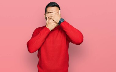 Gündelik kıyafetler giyen genç İspanyol adam gözlerini ve ağzını elleriyle kapatıyor, şaşırmış ve şok olmuş. duygu gizleme 