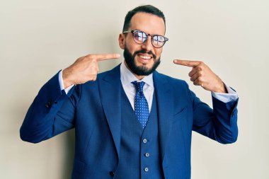 Sakallı, takım elbiseli, kravatlı, neşeli gülümseyen, parmaklarıyla ve ağzıyla işaret eden genç bir adam. diş sağlığı kavramı. 