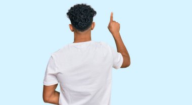 Gündelik beyaz tişört giyen genç Arap adam tersten parmağıyla öne doğru poz veriyor. 