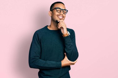 Sıradan kıyafetler ve gözlük takan genç Afro-Amerikalı adam kameraya kendinden emin bakıyor kollarını kavuşturmuş ve çenesini kaldırmış gülümsüyor. Olumlu düşünmek. 