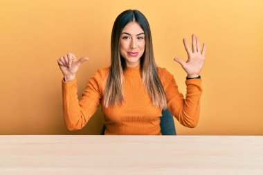 Latin kökenli genç bir kadın, gündelik giysiler içinde masada oturuyor ve 6 numaralı parmaklarıyla işaret ediyor, gülümsüyor ve mutlu.. 