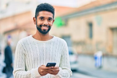 Genç Afrikalı Amerikalı adam şehirde akıllı telefon kullanarak mutlu bir şekilde gülümsüyor..