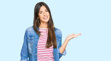 Genç esmer kadın, günlük kıyafetler giyip neşeli bir şekilde gülümsüyor. Avuç içiyle kameraya bakıyor.. 